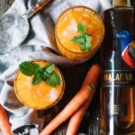 Two orange cocktails garnished with mint next to some young carrots and a bottle of Malabar Spiced Liqueur.
