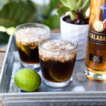 Two Malabar Cuba Libra Cocktails in short glasses next to a lime and bottle of Malabar Spiced Liqueur on a silver metal tray.