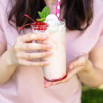 A young woman holds a tall glass filled with a Malabar Strawberry Vanilla Milkshake topped with whipped cream, a strawberry, and a paper straw.