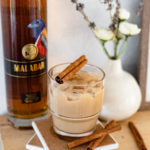 A bottle of Malabar Spiced Liqueur next to an iced rumchata coffee garnished with a cinnamon stick.