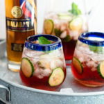 Two short glasses filled with Malabar Rosé Cucumber Cooler garnished with cucumber slices next to a bottle of Malabar Spiced Liqueur.