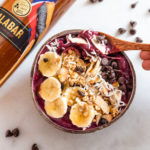 The Malabar Acai Bowl topped with bananas, coconut, and dark chocolate chips next to a bottle of Malabar Spiced Liqueur.