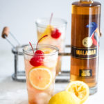 two tom collins cocktails next to a bottle of Malabar Spiced Liqueur and lemons.