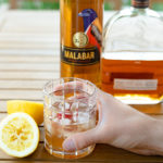 A Malabar Whiskey Sour Cocktail in a short glass next to a lemon and a bottle of Malabar Spiced Liqueur