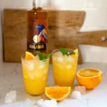 Two Malabar Screwdriver cocktails in short glasses next to orange slices and a bottle of Malabar Spiced Liqueur.