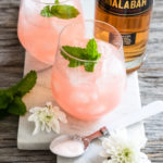 Two small cocktail glasses with Spiced Bubbly Pink Lemonade next to a bottle of Malabar Spiced Liqueur.