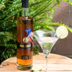 A Malabar Gimlet in a martini glass garnished with lime next to a bottle of Malabar Spiced Liqueur.